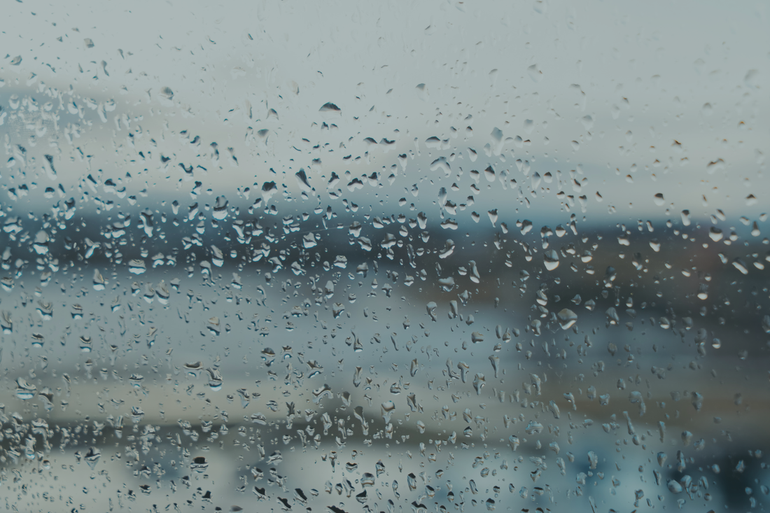 Rain-covered window with a blurred grey landscape beyond, evoking the feeling of being physically present but mentally elsewhere — from a BRÍ Wellness blog post on stress, constant availability, and the hidden cost of always being on.