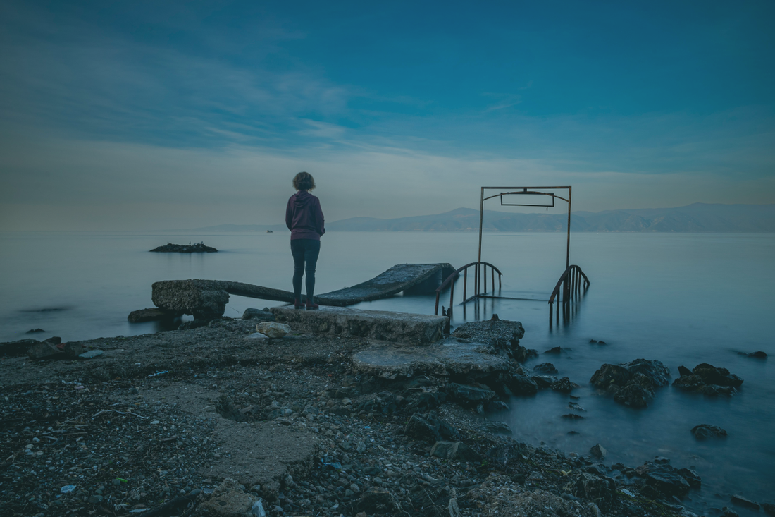 A woman stands alone on a ruined waterside structure in blue pre-dawn light, the still water and half-submerged ironwork around her — from a BRÍ Wellness blog post on a year of disrupted sleep, what helped, and the research behind natural sleep support.