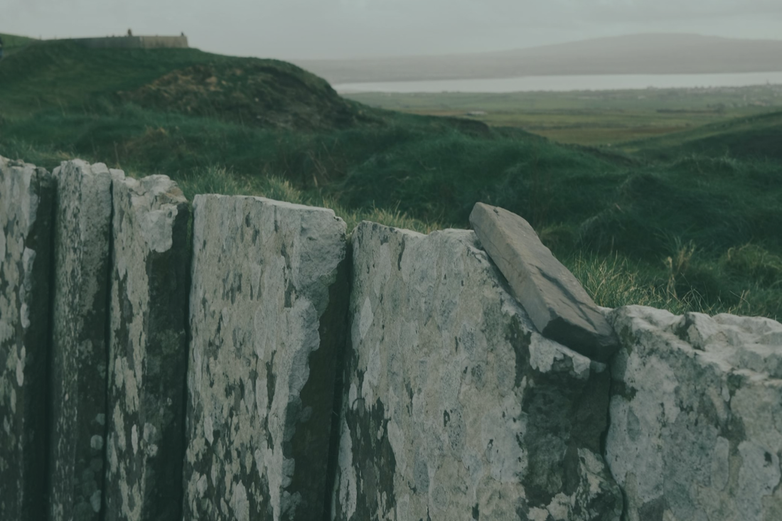 Lichen-covered limestone slabs form an ancient dry-stone wall on the west coast of Ireland, green headland and grey sea stretching to the horizon beyond.